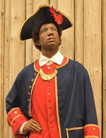 A model of Francisco Menendez, a Black militia member poses with one hand on his hip. He wears a triangular hat and overcoat, both navy with gold trim. His uniform is a white collared shirt with a bright red overshirt.