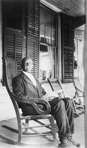 An elderly black man sits in a rocking chair on a porch and gazes into the camera. He is bald with a white mustache. He is wearing a dark suit, white shirt, and leather boots.